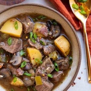 Bowl of steak and potato soup with a gold spoon to the side along with a red napkin and a bowl of chopped green onions.