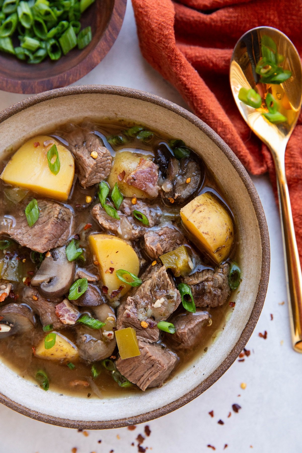 Bowl of steak and potato soup with a gold spoon to the side along with a red napkin and a bowl of chopped green onions.