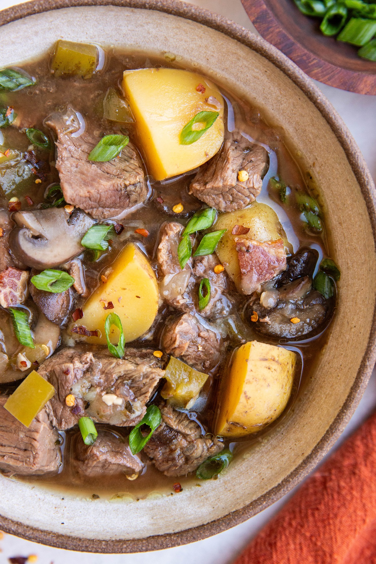 Big bowl of steak and potato soup with a bowl of chopped green onions and a red napkin to the side.