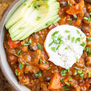 Large bowl of pumpkin chili with sliced avocado, chopped chives, and sour cream on top.