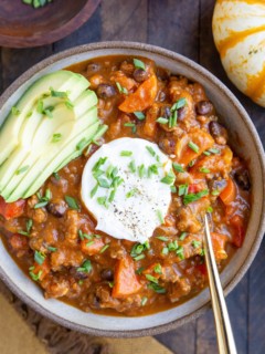 Bowl of beef pumpkin chili on a wood background with avocado, sour cream, chives, and a gold spoon.