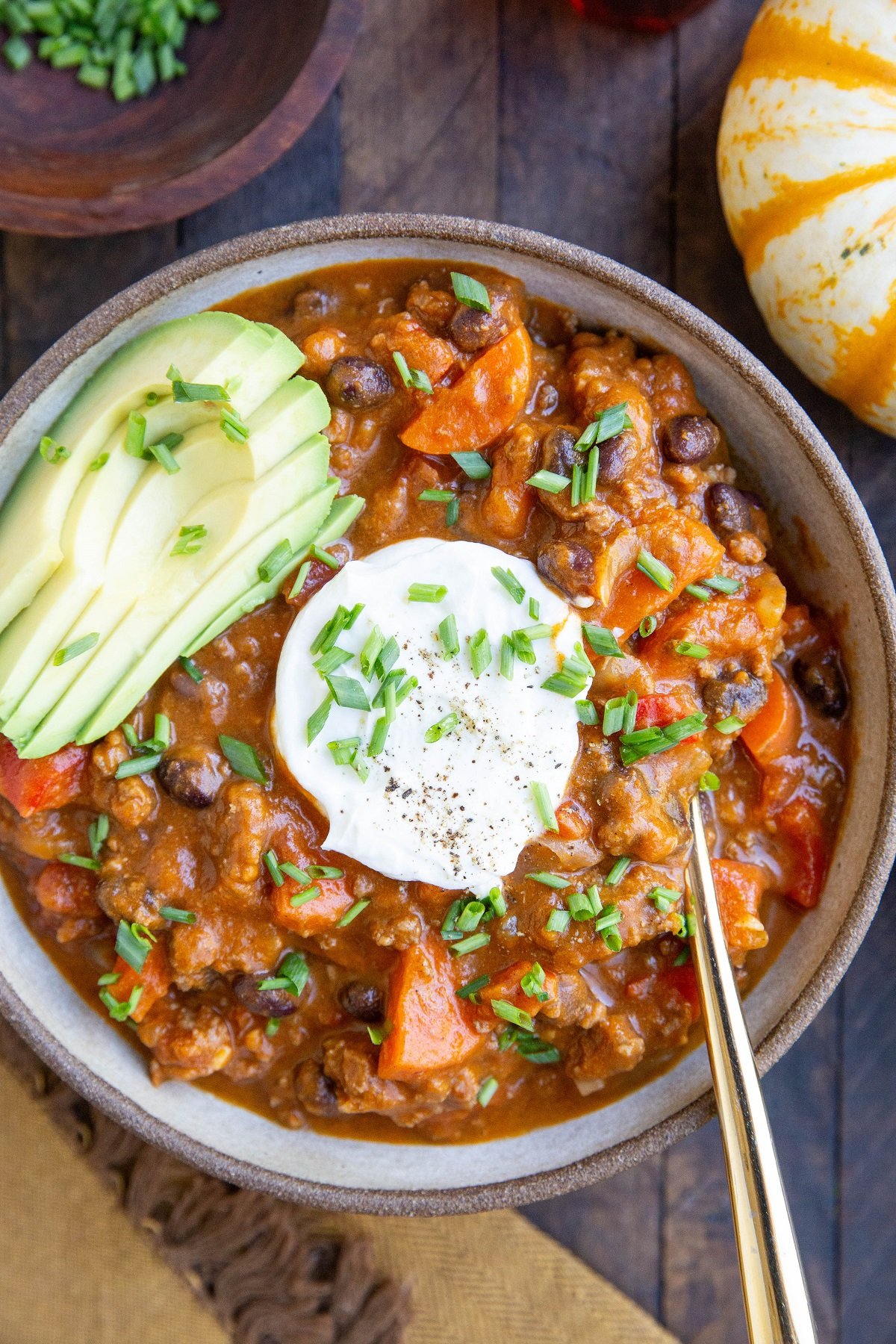 Bowl of beef pumpkin chili on a wood background with avocado, sour cream, chives, and a gold spoon.