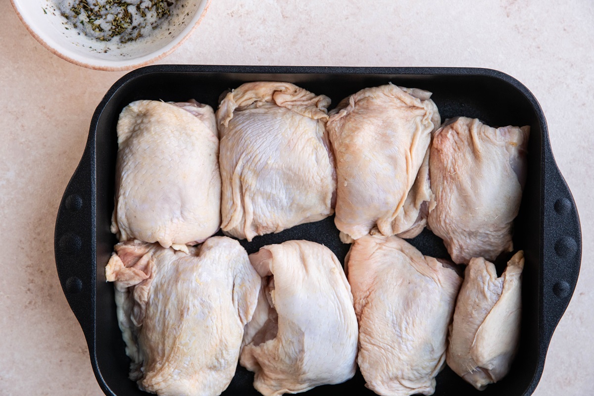 Raw bone-in skin-on chicken thighs in a large casserole dish with a bowl of garlic butter sauce to the side.