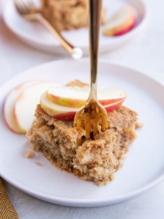 Slice of apple cake on a white plate with a fork taking a bite.