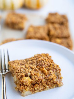 A slice of pumpkin apple oatmeal bar on a white plate with a gold fork and more bars in the background.