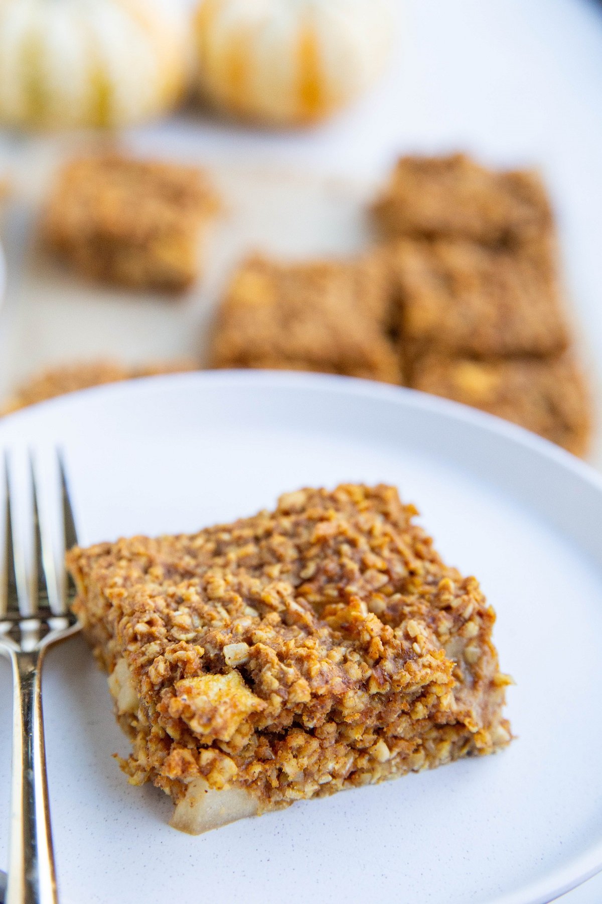 A slice of pumpkin apple oatmeal bar on a white plate with a gold fork and more bars in the background.