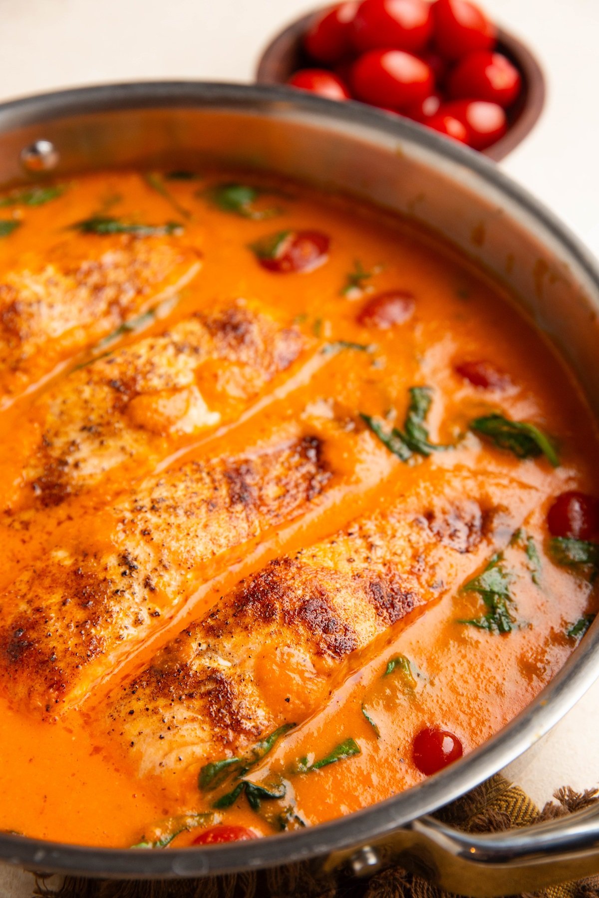 Stainless steel skillet with four cooked salmon filets in creamy red pepper sauce and fresh tomatoes in the background.