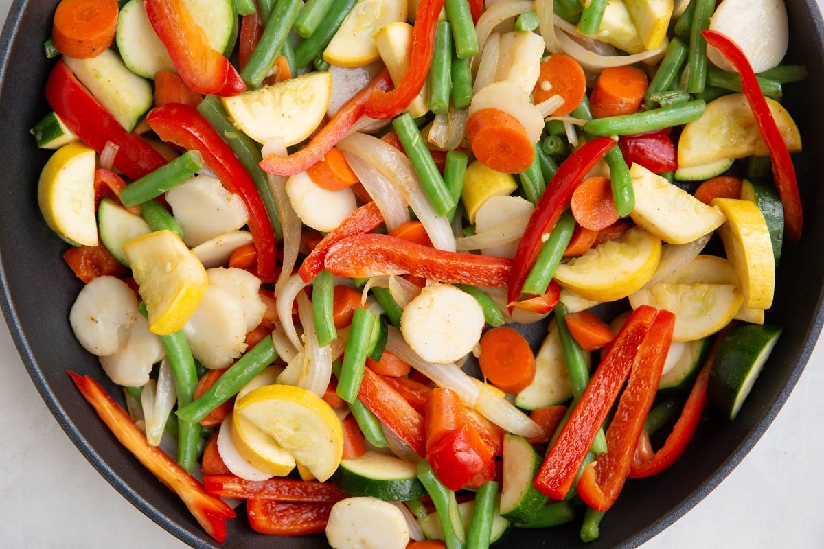 Onion, bell pepper. green beans, zucchini, yellow squash, carrots and water chestnuts cooking in a skillet.