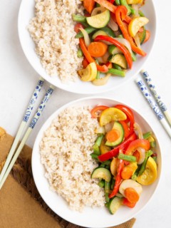 Two white bowls of stir-fry vegetables with brown rice and chopsticks.