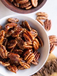 White bowl of candied pecans with raw pecans all around.