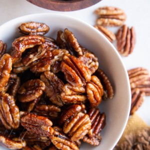 White bowl of candied pecans with raw pecans all around.