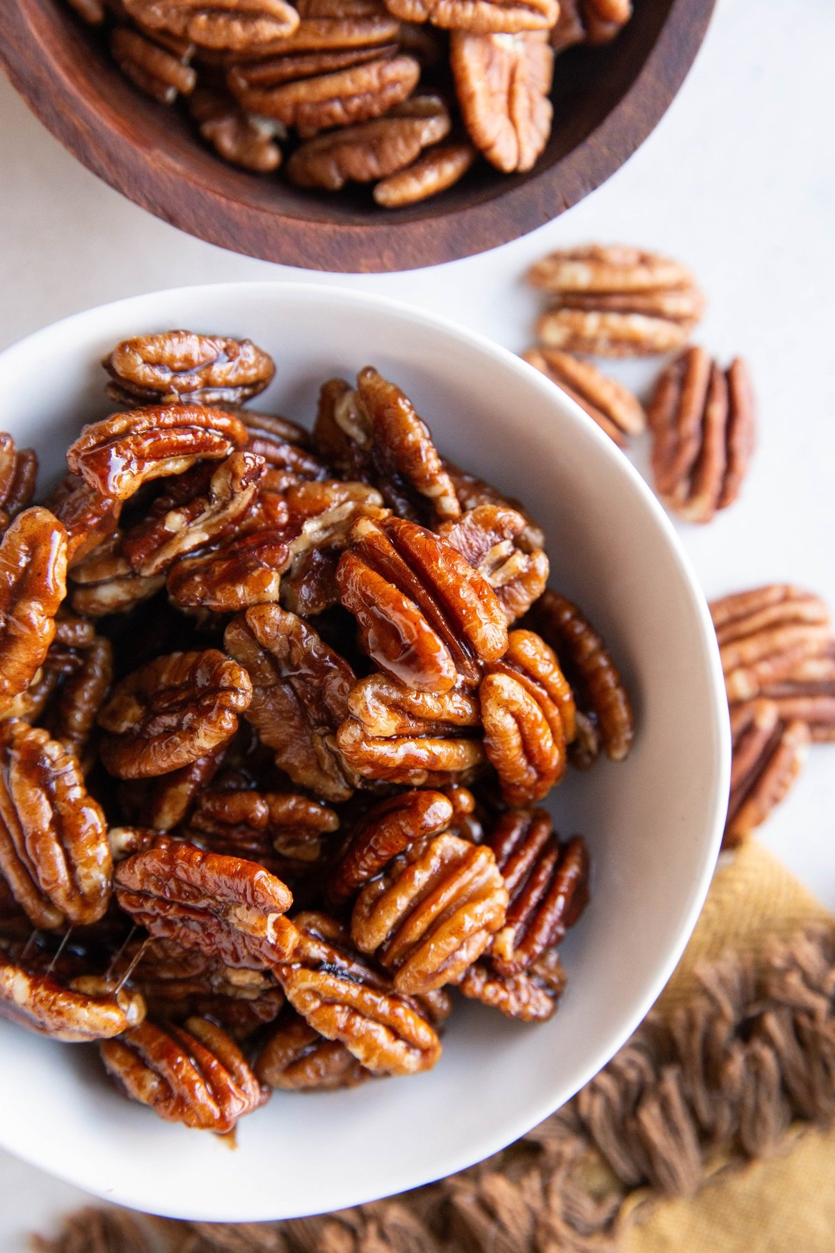 White bowl of candied pecans with raw pecans all around.
