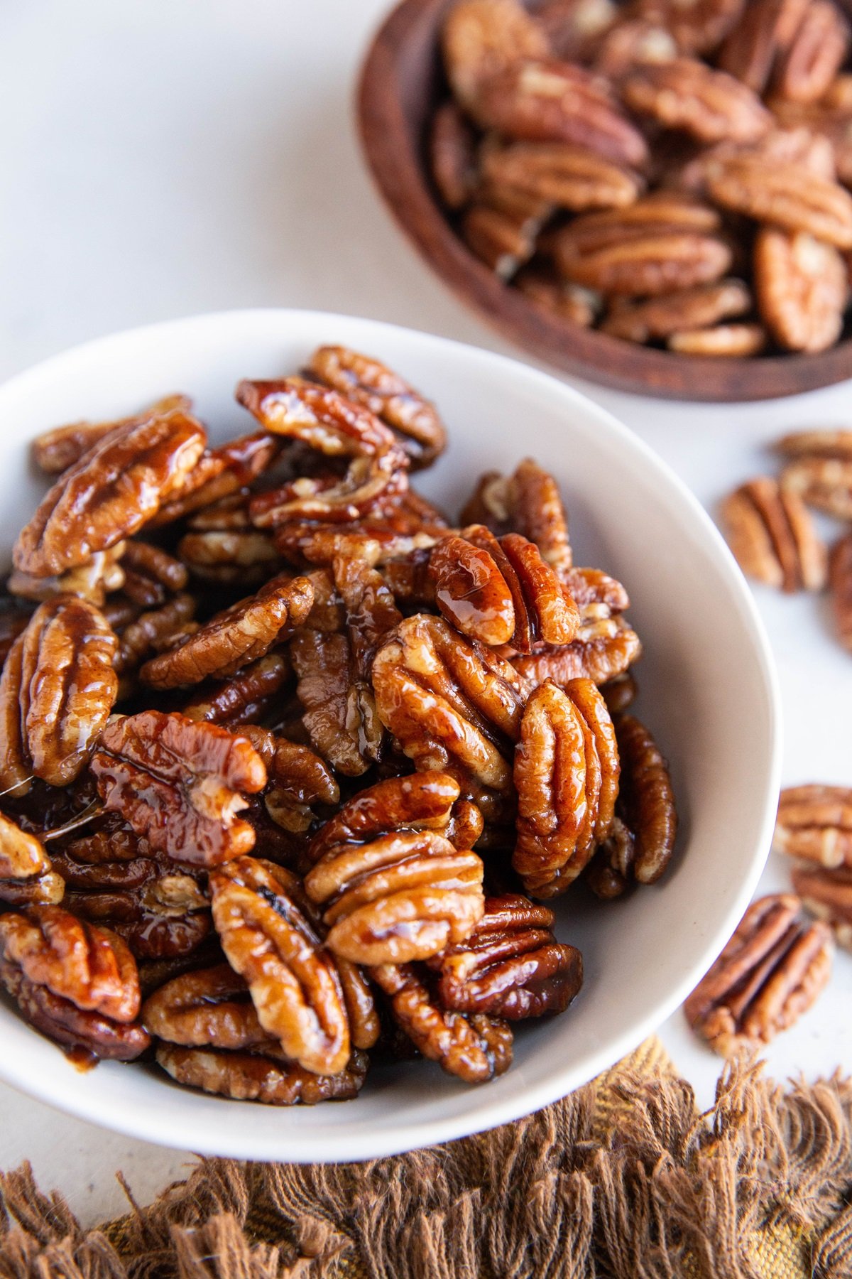 White bowl of maple candied pecans. A bowl of raw pecans in the background.