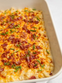 White casserole dish with baked creamy cheesy cauliflower sprinkled with bacon and chives.