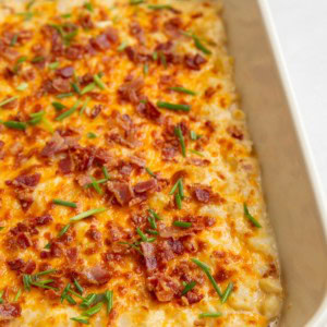 White casserole dish with baked creamy cheesy cauliflower sprinkled with bacon and chives.