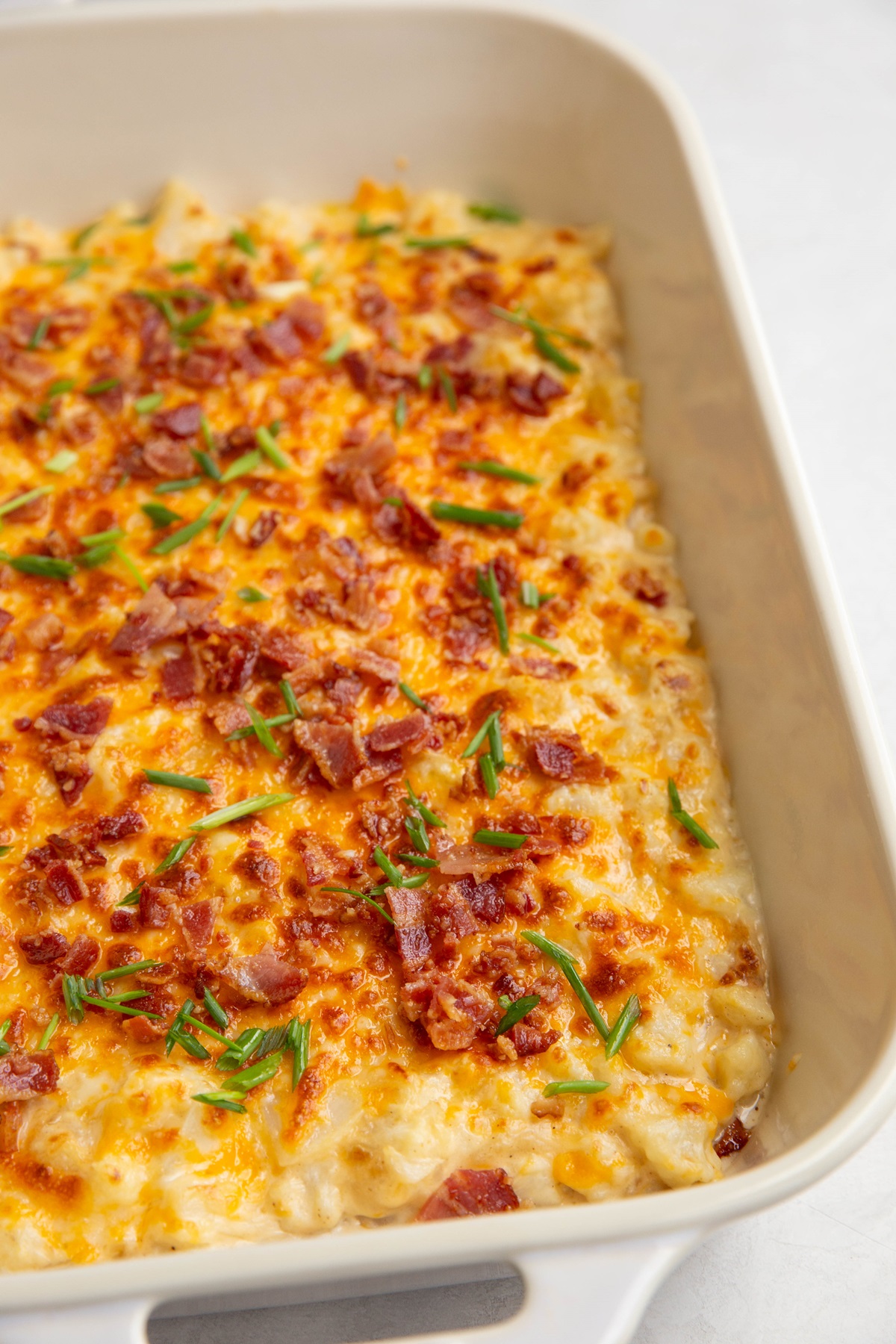 White casserole dish with baked creamy cheesy cauliflower sprinkled with bacon and chives.