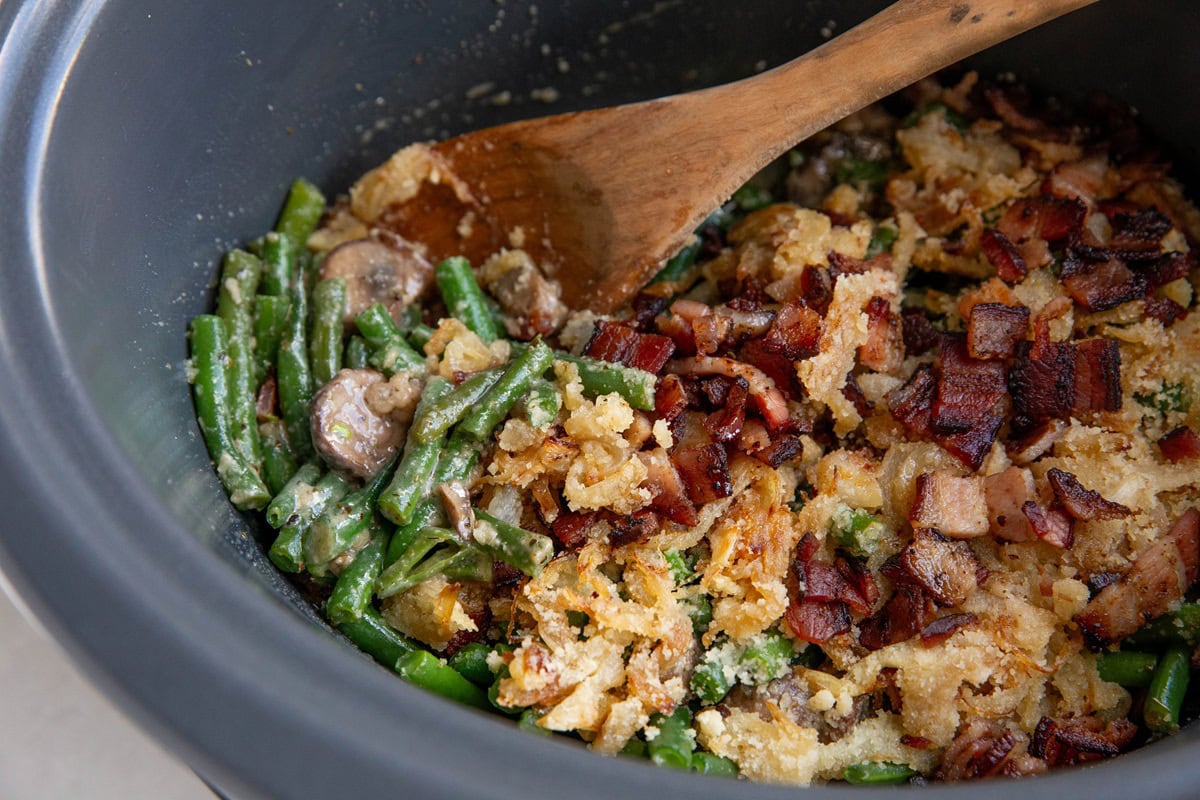 green bean casserole in a slow cooker with a wooden spoon, in the process of being served.