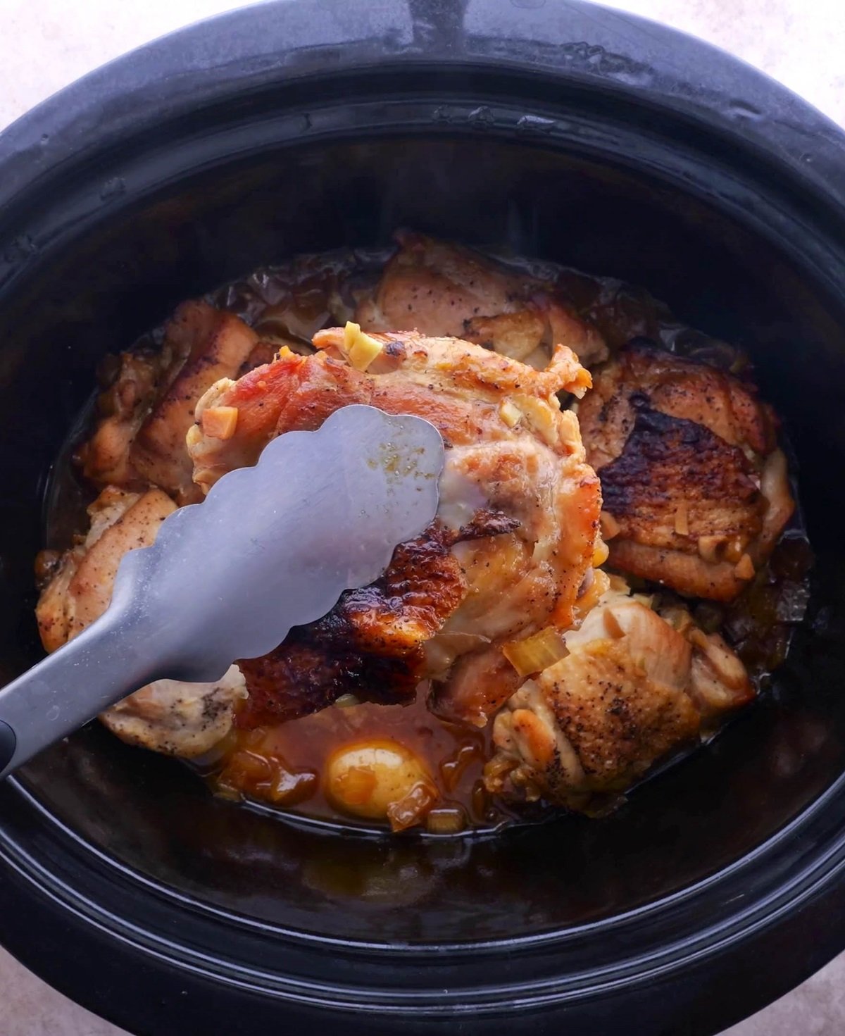 Tongs holding a chicken thigh out of a crock pot, finished cooking.