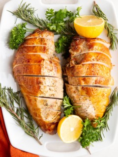 Garlic herb turkey breasts on a white serving platter surrounded by fresh herbs and lemon.