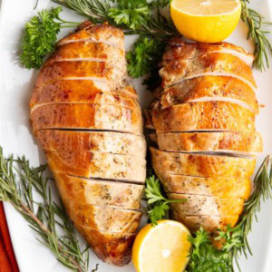 Garlic herb turkey breasts on a white serving platter surrounded by fresh herbs and lemon.