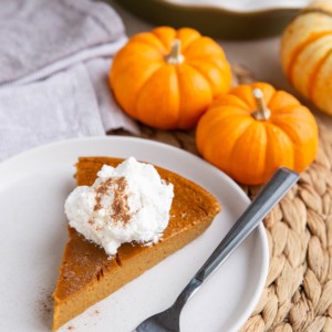 White plate with a slice of crustless pumpkin pie with whipped cream on top and the rest of the pie in the background.