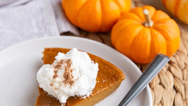 White plate with a slice of crustless pumpkin pie with whipped cream on top and the rest of the pie in the background.