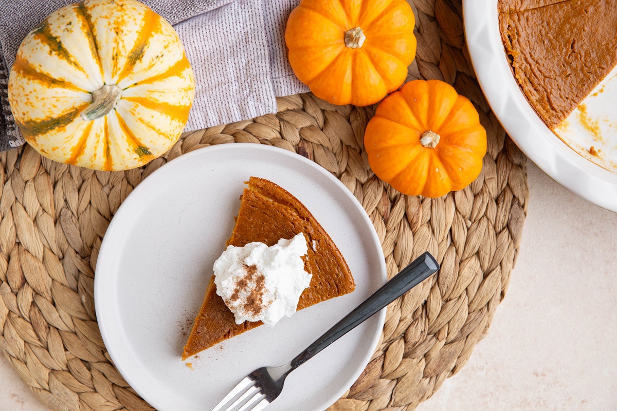 Plate with a slice of pumpkin pie on a place mat with the rest of the pumpkin pie to the side and fresh pumpkins around.