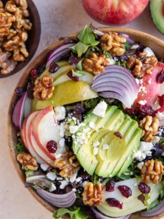 Wooden bowl of salad with avocado, apples, pears and more. A bowl of candied walnuts to the side.
