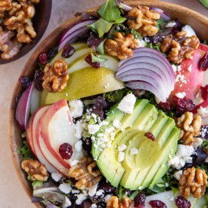 Wooden bowl of salad with avocado, apples, pears and more. A bowl of candied walnuts to the side.
