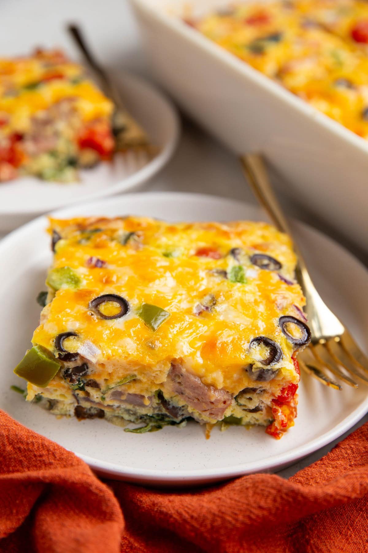 Large slice of egg casserole on a white plate with another slice in the background and a casserole dish.