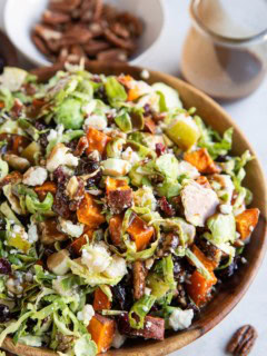 Bowl of shaved brussel sprout salad with all sorts of goodies inside and balsamic vinaigrette in the background.