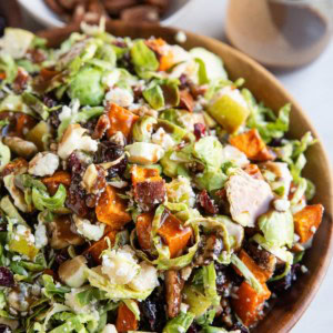 Bowl of shaved brussel sprout salad with all sorts of goodies inside and balsamic vinaigrette in the background.