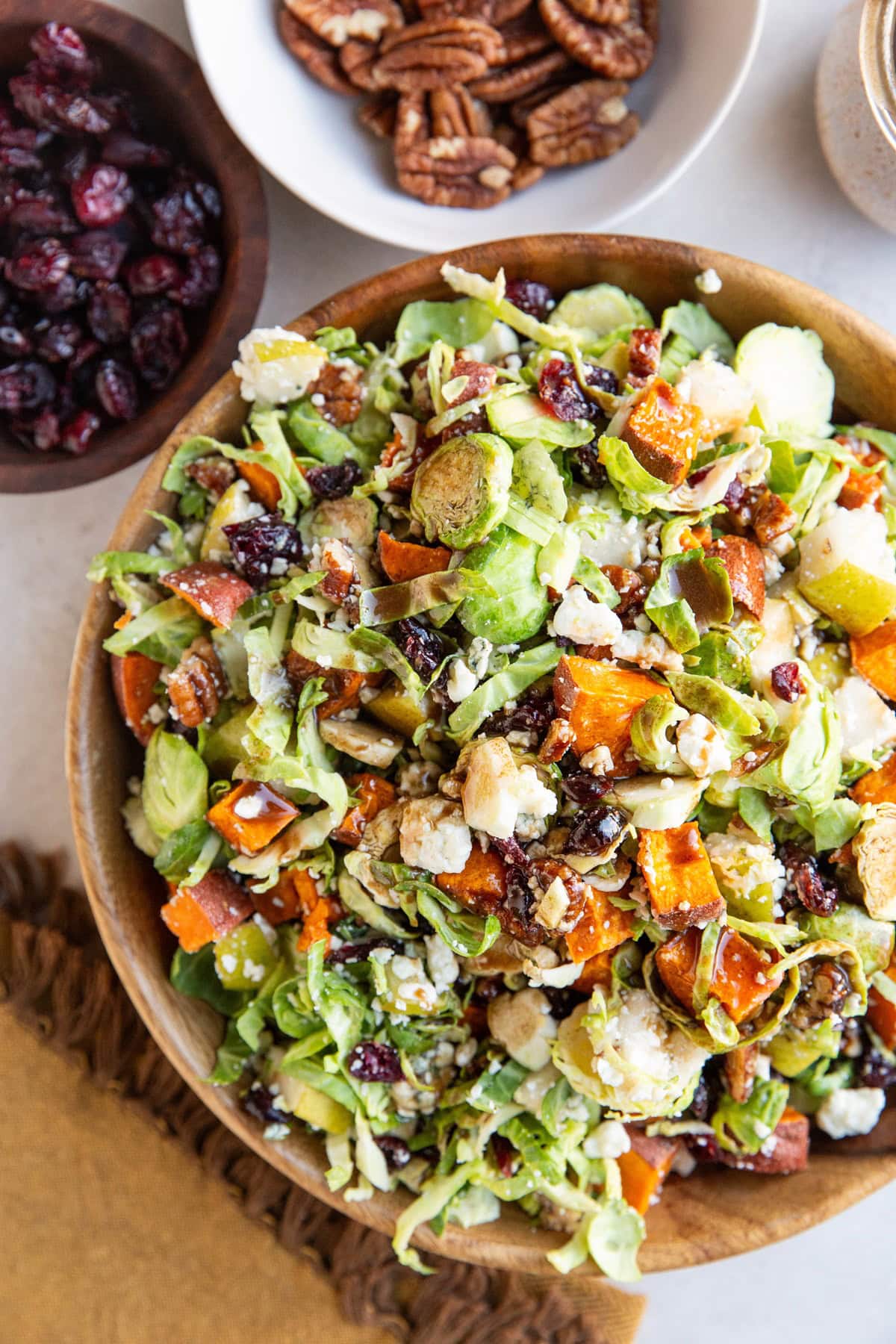 Bowl of shaved brussel sprout salad with roasted sweet potatoes and bowls of dried cranberries and pecans to the side with a golden napkin.