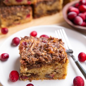 Cranberry orange coffee cake on a white plate with slices of cake in the background and fresh cranberries all around.