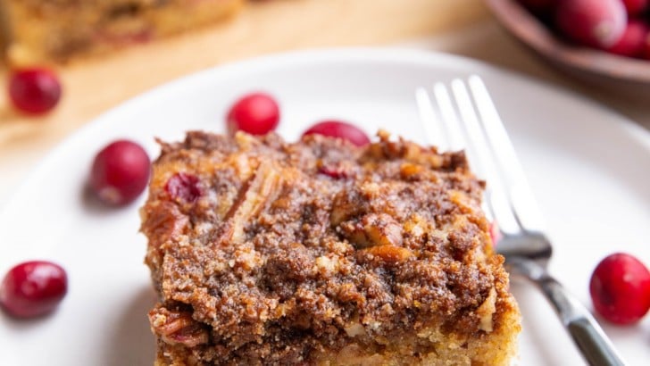 Cranberry orange coffee cake on a white plate with slices of cake in the background and fresh cranberries all around.