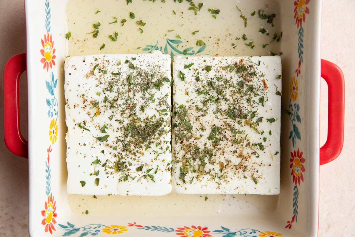 Two blocks of feta cheese in a baking dish with avocado oil and dried herbs.