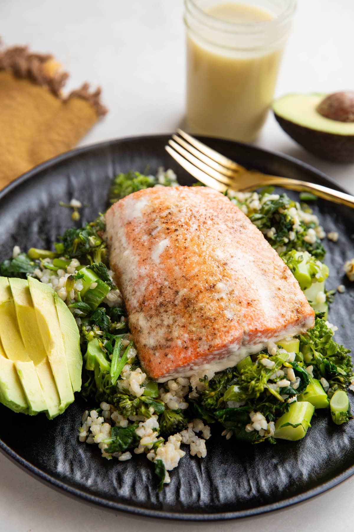 Baked sockeye salmon on top of rice and veggies with a side of avocado on a plate with dressing in the background.