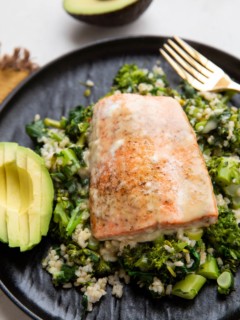 Sockeye salmon on a black plate with rice, broccoli, and spinach with sliced avocado to the side.
