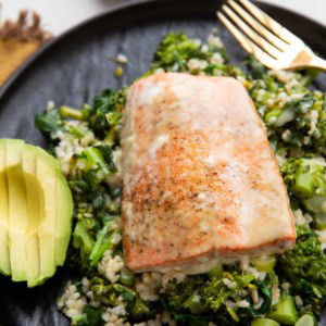 Sockeye salmon on a black plate with rice, broccoli, and spinach with sliced avocado to the side.