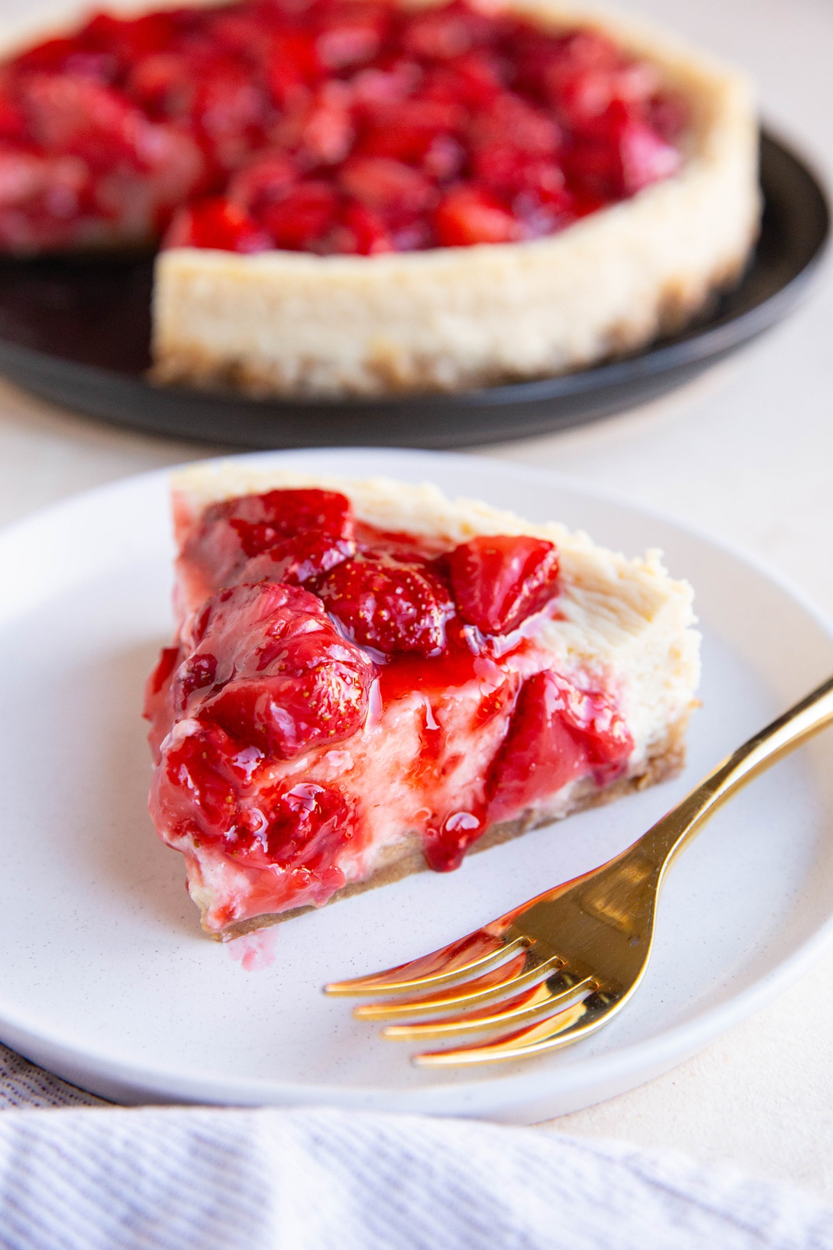 Slice of cottage cheese cheesecake on a white plate with strawberry topping and the rest of the cheesecake in the background.