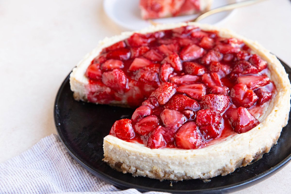 Cottage cheese cheesecake on a black plate topped with strawberry compote with a slice taken out of the cheesecake.