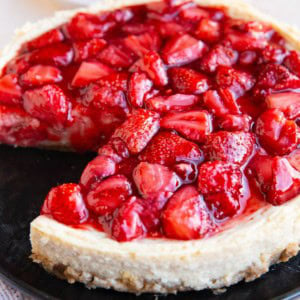 Cheesecake on a black plate with strawberry topping and a slice of cheesecake in the background.