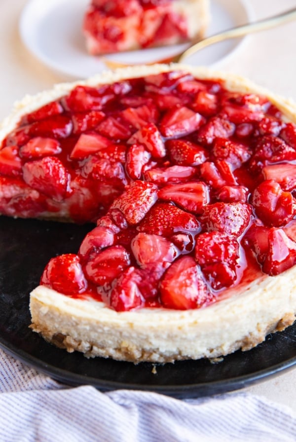 Cheesecake on a black plate with strawberry topping and a slice of cheesecake in the background.