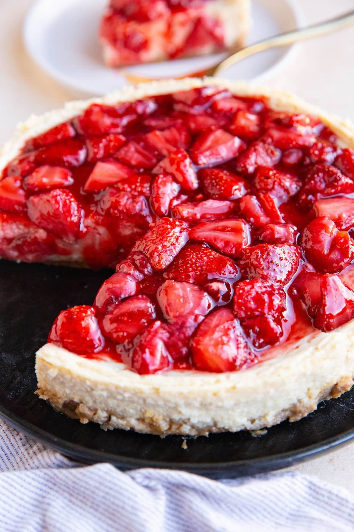 Cheesecake on a black plate with strawberry topping and a slice of cheesecake in the background.