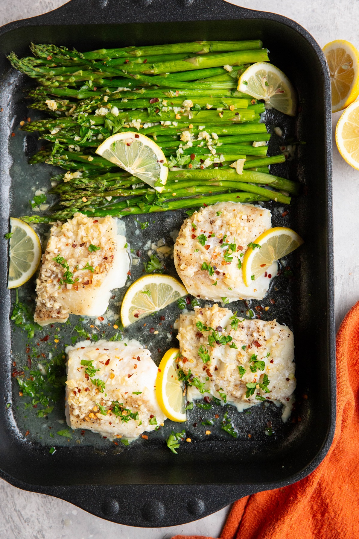 Garlic butter cod in a casserole dish with roasted asparagus and lemon slices. A red napkin to the side.
