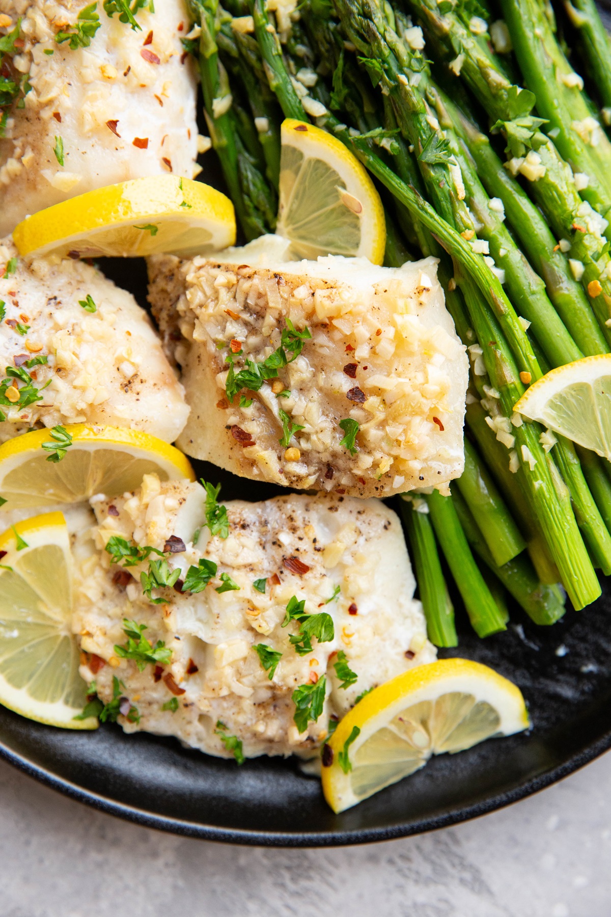 Four baked cod filets on a black plate with baked asparagus and lemon wedges.