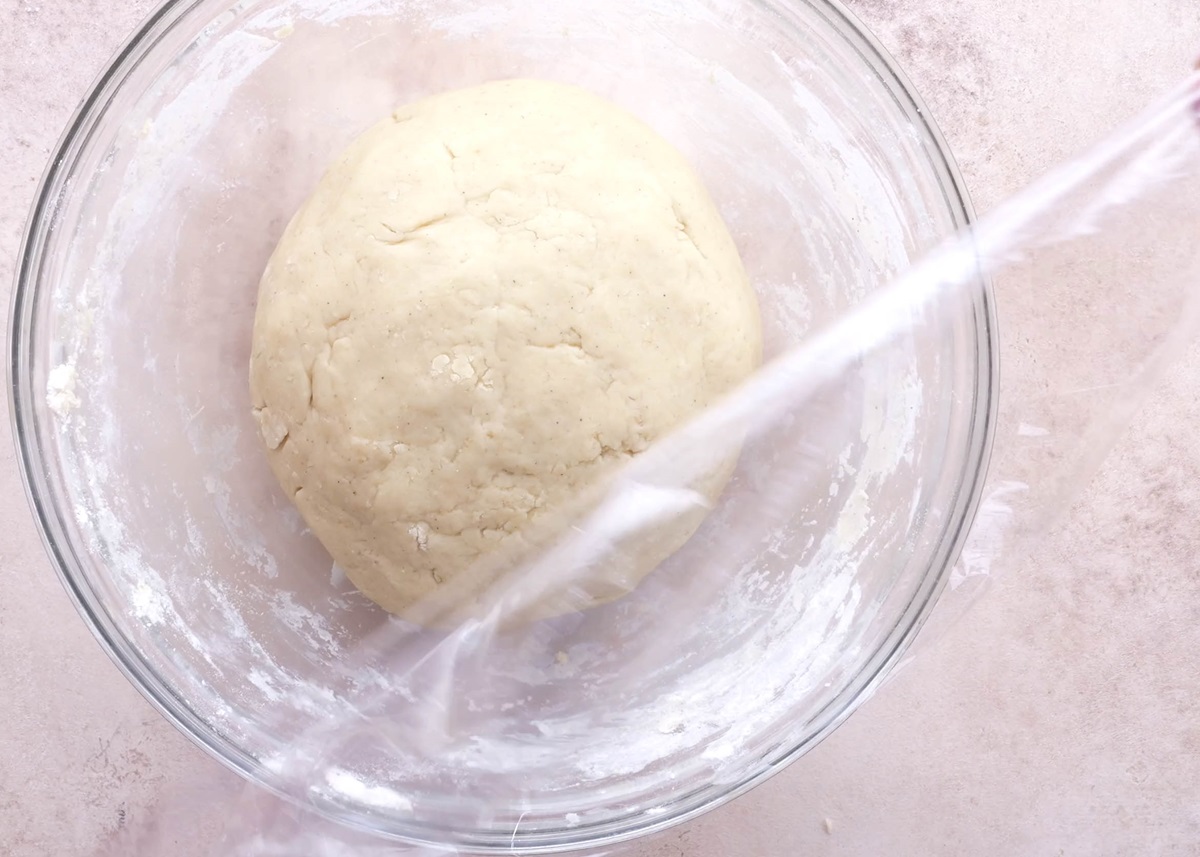 Cinnamon roll dough in a mixing bowl being covered with plastic wrap.