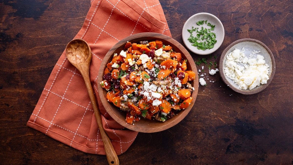 Wooden background with wood bowl of roasted carrots and a bowl of feta and parsley to the side.