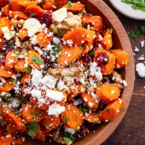 Maple Roasted Carrots in a wooden bowl with feta, dried cranberries and walnuts and a small bowl of parsley to the side.