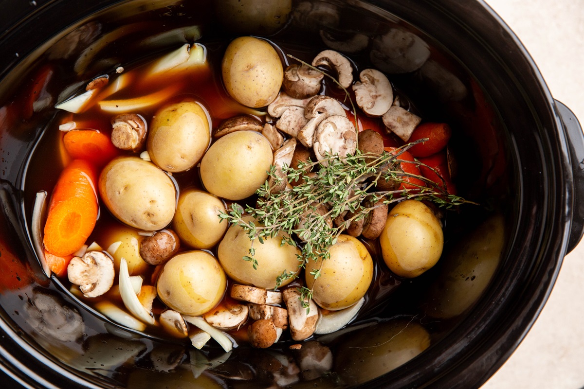 Vegetables, broth, and fresh herbs at the bottom of a crock pot to be cooked with pork roast.